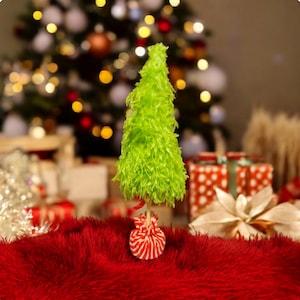 May include: A small, bright green Christmas tree decoration with a textured, feathery appearance. The tree is mounted on a wooden stick and sits atop a red and white striped base. The background features a decorated Christmas tree and wrapped gifts.