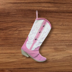 May include: Pink cowboy boot stocking with white embroidery and silver buttons. The stocking is hanging on a wood background.