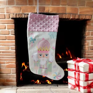 May include: A pink and white Christmas stocking with a nutcracker design. The stocking has a pink, fuzzy cuff and a white background with a nutcracker illustration. The nutcracker is wearing a green and purple uniform and a gold crown. The stocking is hanging in front of a fireplace.