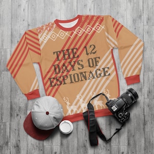 May include: A tan sweatshirt with red and white stripes and the text "THE 12 DAYS OF ESPIONAGE." The sweatshirt has a red collar and cuffs, with a white and red pattern on the shoulders. A gray and maroon baseball cap, a camera, and a coffee cup are also in the image.