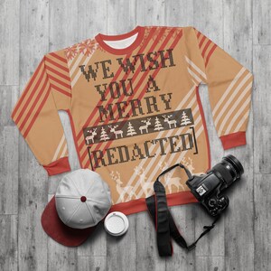 Może przedstawiać: Beżowy sweter świąteczny z napisem "WE WISH YOU A MERRY [REDACTED]" w kolorze czarnym. Sweter ma czerwono-białe paski i wzór reniferów i drzew. Widoczna jest również biało-bordowa czapka z daszkiem, aparat fotograficzny i filiżanka do kawy.