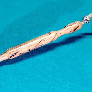 May include: A wooden stirring stick with a decorative knob on one end. The stick is made of light brown wood with dark brown streaks. The knob is made of a darker wood and has a silver metal accent.