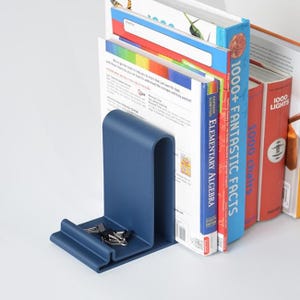 Op de afbeelding: Een marineblauwe metalen boekensteun met een gebogen ontwerp, die verschillende boeken van verschillende formaten en kleuren vasthoudt. De boekensteun heeft een klein bakje aan de basis, met paperclips. De boeken bevatten titels als "1000+ Fantastic Facts" en "Elementary Algebra".