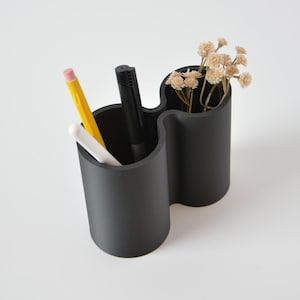 May include: Black double-sided desk organiser with a wavy design. It holds pens, pencils, and a small bouquet of dried flowers.