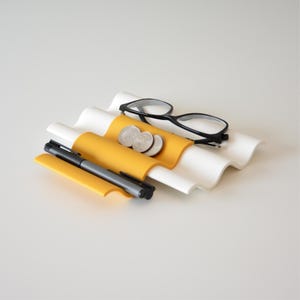 May include: A white and yellow wavy desk organizer holds a pair of black-framed glasses, a pen, and three coins. The organizer has a modern design and is made of a combination of white and yellow materials.