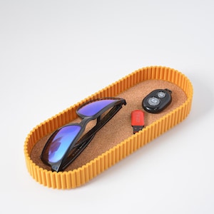 Puede incluir: Una bandeja oblonga amarilla con base de corcho contiene gafas de sol negras, un mando a distancia negro y una unidad USB roja. La bandeja tiene un borde acanalado. Las gafas de sol tienen lentes reflectantes azules.