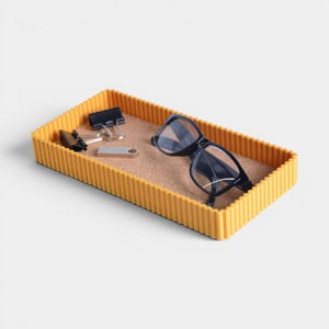 May include: A rectangular mustard yellow tray with a ribbed exterior and a cork base. Inside the tray are a pair of black-framed eyeglasses, two black metal binder clips, and a small silver-colored rectangular USB flash drive.