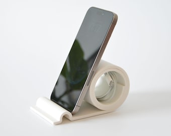 Puede incluir: Un soporte para teléfono blanco y beige con un teléfono inteligente encima. El soporte tiene un diseño curvo con una ranura para sostener el teléfono en posición vertical. La pantalla del teléfono muestra la hora 11:17.