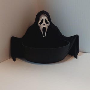 Puede incluir: Un cuenco negro con una máscara blanca de Ghostface con una boca larga y distorsionada y ojos huecos, unida a una capucha de tela negra que cae sobre los lados del cuenco, creando una pieza de exhibición oscura y dramática para guardar objetos.