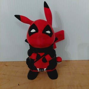 May include: A red and black figurine of a Pikachu character in a Deadpool costume. The figure has black markings around the eyes and a black outfit. The ears and tail are red with black tips. The figure is standing on a wooden surface.