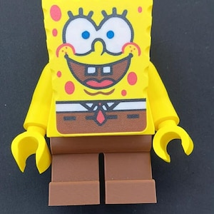 Puede incluir: Una minifigura amarilla de Bob Esponja con cabeza rectangular, grandes ojos azules, manchas rosas y una amplia sonrisa. El torso muestra una camisa blanca con corbata roja y las piernas son de plástico de color marrón sólido.