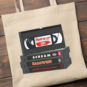 May include: A beige tote bag featuring a stack of VHS tapes. The top tape reads "Friday the 13th VHS" with a blood splatter design. Below are tapes labeled "SCREAM", "HALLOWEEN", and "A Nightmare on Elm Street".
