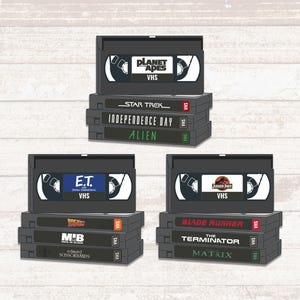 Puede incluir: Una colección de cintas VHS retro con títulos de películas como "Planet of the Apes", "Star Trek", "Independence Day", "Alien", "E.T.", "Blade Runner" y "The Terminator". Las cintas están apiladas.