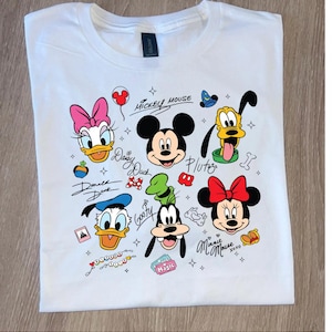 May include: White t-shirt with a colourful graphic of Disney characters, including Mickey Mouse, Minnie Mouse, Donald Duck, Daisy Duck, Goofy, and Pluto. The design includes character names and related icons.