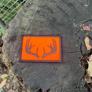 May include: Rectangular patch with an orange background and a dark gray antler design. The patch has a brown border. The patch is placed on a dark gray tree stump.