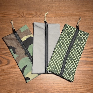 May include: Three zippered pouches with different camouflage patterns. The pouches are made of fabric and have black zippers. The pouches are laid out on a wooden surface.