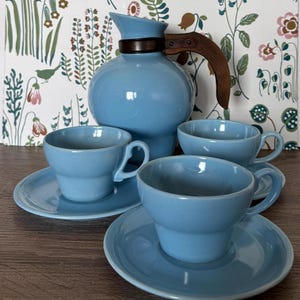May include: A light blue ceramic coffee set featuring a round pitcher with a wooden handle and four matching cups and saucers. The set is displayed on a wooden surface with a floral patterned backdrop.