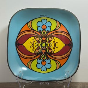 May include: A square, turquoise plate with a black rim and a colorful, symmetrical design. The design features orange, yellow, and maroon shapes with two blue flower motifs. The plate has the artist's signature in the upper right corner.