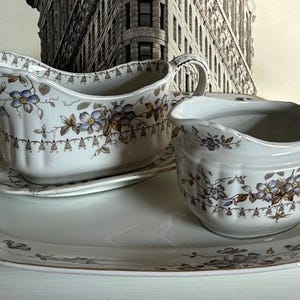 May include: A white porcelain gravy boat with a floral design and a matching saucer. The gravy boat has a handle and a spout. The saucer has a scalloped edge. The set is on a larger white porcelain serving platter with a floral design.