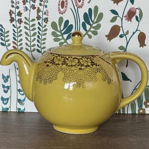 May include: Yellow ceramic teapot with gold floral detailing around the rim and on the lid. The teapot has a curved spout and handle, and a gold knob on the lid. The background features a floral wallpaper.