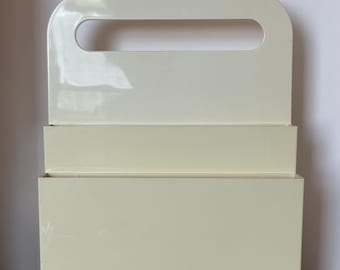 May include: A cream-colored, rectangular organizer with a handle. The organizer has a box-like bottom section and a smaller, raised section above it. The handle is a curved opening at the top, and the overall design is minimalist.