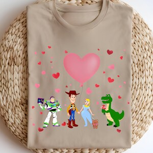 May include: A tan t-shirt featuring a graphic of cartoon characters, including Buzz Lightyear, Woody, and a princess, holding hands under a large pink heart balloon. Scattered pink and red hearts decorate the background.