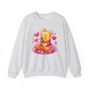Puede incluir: Una sudadera gris con una imagen de dibujos animados de Winnie de Pooh sosteniendo un coraz&oacute;n rodeado de corazones rosas y flores.