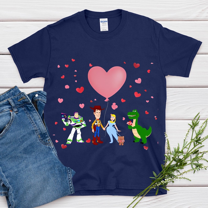 May include: Navy blue t-shirt featuring cartoon characters Buzz Lightyear, Woody, a princess, and a green dinosaur. A large pink heart balloon floats above them, with scattered red and pink hearts. The shirt is paired with blue jeans.
