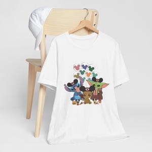Puede incluir: Camiseta blanca con un gr&aacute;fico de Stitch, Baby Yoda y Groot con orejas de Mickey Mouse y sosteniendo bocadillos. Los personajes est&aacute;n rodeados de globos de Mickey Mouse de colores.