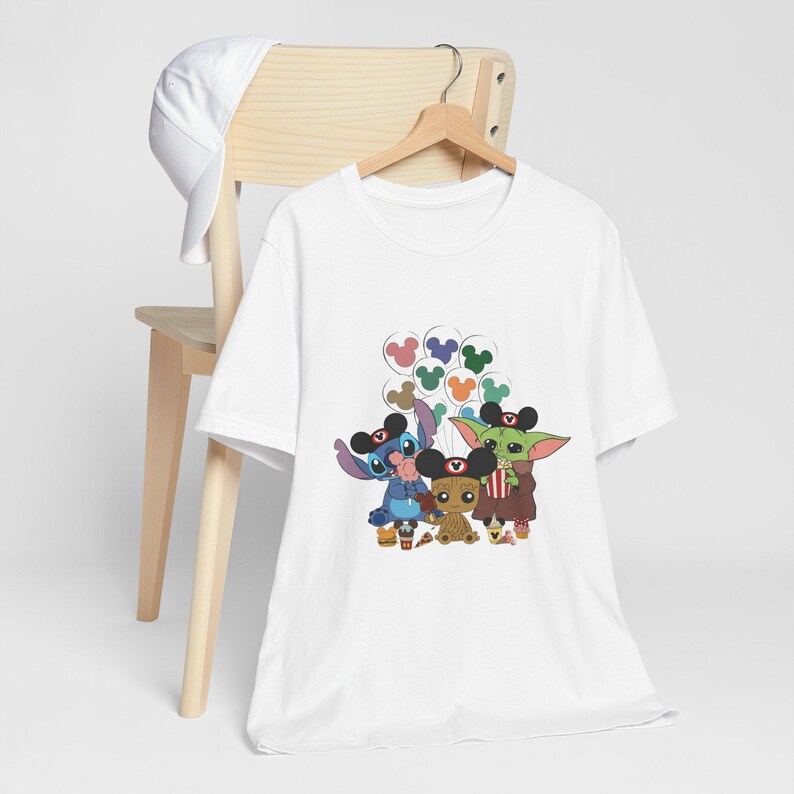 Puede incluir: Camiseta blanca con un gr&aacute;fico de Stitch, Baby Yoda y Groot con orejas de Mickey Mouse y sosteniendo bocadillos. Los personajes est&aacute;n rodeados de globos de Mickey Mouse de colores.