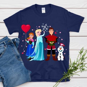 Pu&ograve; includere: T-shirt blu navy con una grafica a cartoni animati di personaggi di un popolare film d'animazione. Il design include un palloncino a forma di cuore rosso, un fiocco di neve e un bouquet di rose rosse. La maglietta &egrave; mostrata con un paio di jeans.