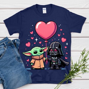 May include: Navy blue t-shirt with a cartoon illustration of Baby Yoda and Darth Vader holding hands and a pink heart-shaped balloon. Small pink hearts and stars are scattered around the characters. The shirt is paired with blue jeans.