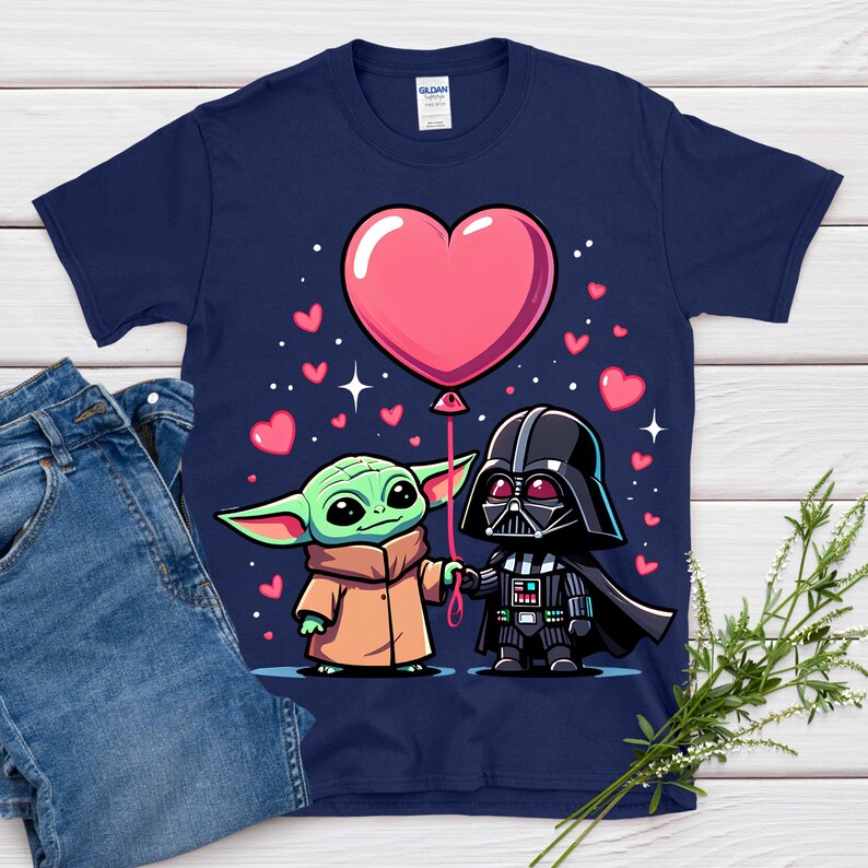 May include: Navy blue t-shirt with a cartoon illustration of Baby Yoda and Darth Vader holding hands and a pink heart-shaped balloon. Small pink hearts and stars are scattered around the characters. The shirt is paired with blue jeans.