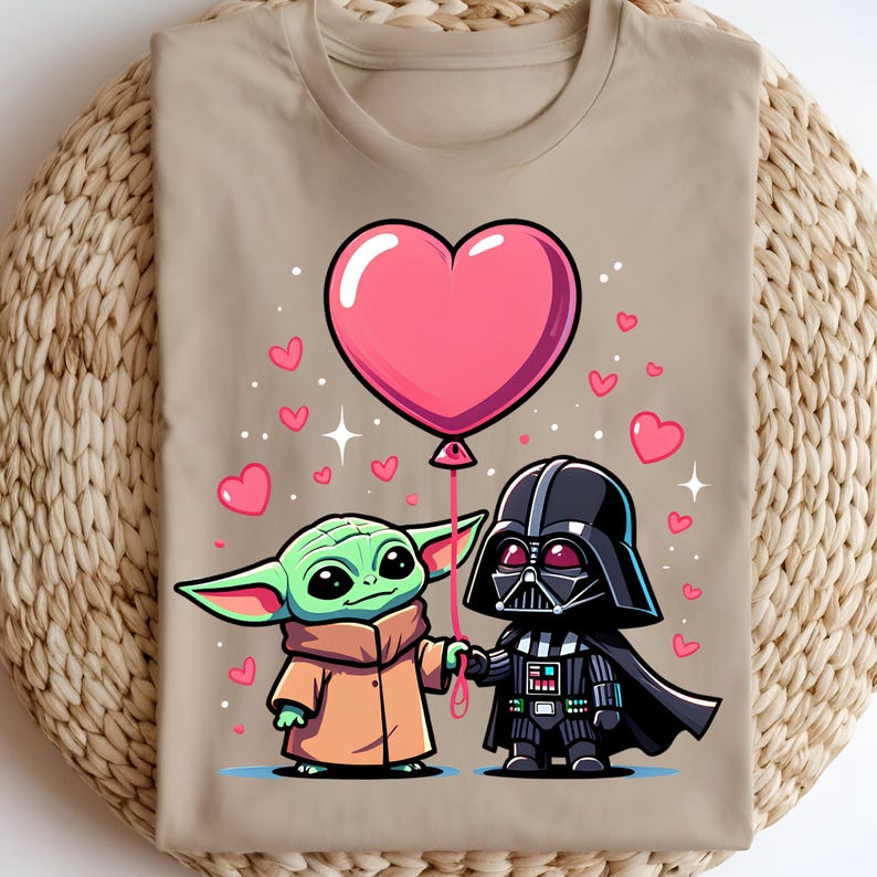 May include: Beige t-shirt with a cartoon illustration of Baby Yoda and Darth Vader holding a pink heart-shaped balloon. Small pink hearts and stars are scattered around the characters. The design is centered on the shirt.