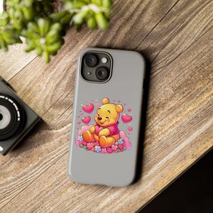 Puede incluir: Una funda de tel&eacute;fono gris con una imagen de dibujos animados de Winnie de Pooh sosteniendo un coraz&oacute;n. El fondo es gris claro con corazones y flores rosas.