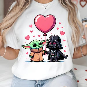 May include: White t-shirt with a cartoon illustration of Grogu and Darth Vader holding hands, with a pink heart-shaped balloon. Small pink hearts are scattered around the characters. The characters are in a cartoon style.