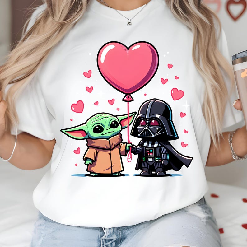 May include: White t-shirt with a cartoon illustration of Grogu and Darth Vader holding hands, with a pink heart-shaped balloon. Small pink hearts are scattered around the characters. The characters are in a cartoon style.
