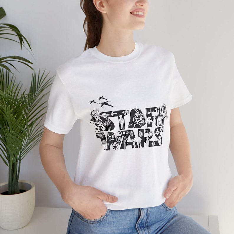 Może przedstawiać: Biała koszulka z czarno-białym nadrukiem logo Star Wars, złożonym z r&oacute;żnych postaci i przedmiot&oacute;w ze świata Gwiezdnych Wojen.