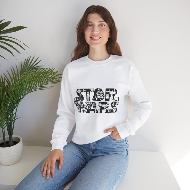 K&ouml;nnte beinhalten: Wei&szlig;er Pullover mit einem schwarzen Aufdruck des Star Wars-Logos, das aus verschiedenen Figuren und Symbolen besteht.