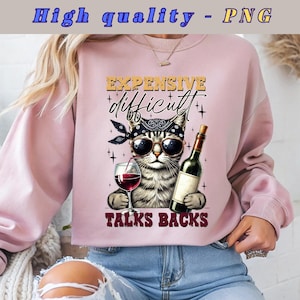 May include: Pink sweatshirt featuring a graphic of a cat wearing sunglasses and a bandana, holding a glass of wine and a bottle of wine. The text on the sweatshirt reads "Expensive, difficult, talks backs".