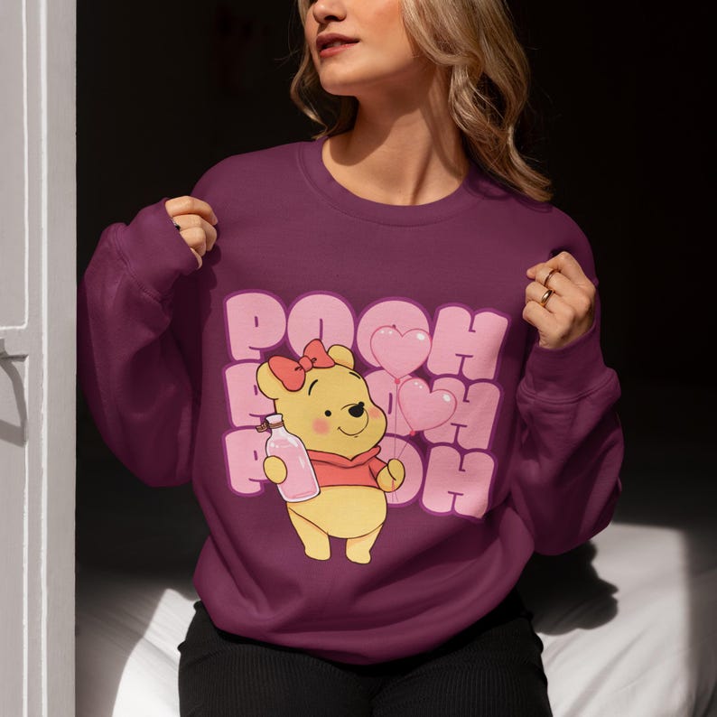 May include: A maroon sweatshirt with a cartoon image of Winnie the Pooh holding a bottle and pink balloons. The text "POOH" is repeated in a pink, bubble-like font.