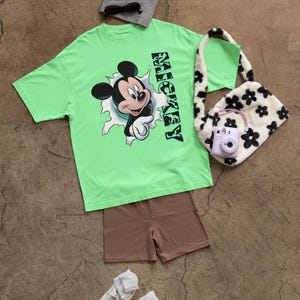 以下が含まれることがあります： 穴から顔を出しているミッキーマウスのグラフィックがプリントされた緑色のTシャツ。Tシャツの右側に「ミッキー」という文字が縦にプリントされています。Tシャツは、茶色のショートパンツ、白いスニーカー、灰色のビーニー、ピンクのカメラが付いた白黒のふわふわのバッグと組み合わせています。