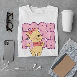 May include: White t-shirt with a cartoon image of Winnie the Pooh holding a bottle of honey and pink heart balloons. The text "POOH" is repeated in pink letters around the image.