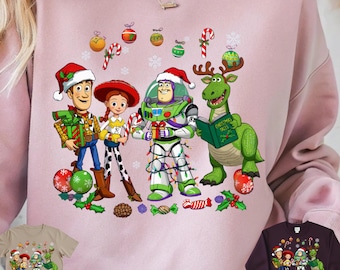 May include: Pink sweatshirt featuring a festive graphic of Toy Story characters Woody, Jessie, Buzz Lightyear, and a green dinosaur, all wearing Santa hats and holding gifts. Christmas ornaments and candy canes decorate the design. The words "Digital Download-PNG File" are at the bottom.
