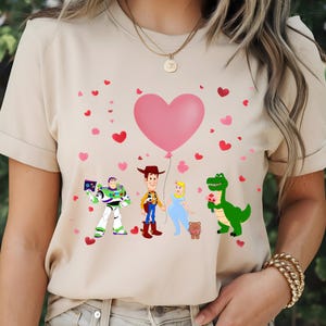 May include: Beige t-shirt featuring cartoon characters Buzz Lightyear, Woody, Cinderella, and Rex, with a pink heart balloon and scattered hearts. The characters are holding hands and flowers, creating a Valentine's Day theme.