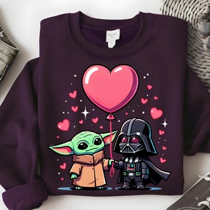 May include: A dark purple sweatshirt with a cartoon illustration of Baby Yoda and Darth Vader holding hands. A large pink heart balloon floats above them, surrounded by smaller pink hearts and stars. The sweatshirt is a solid colour.