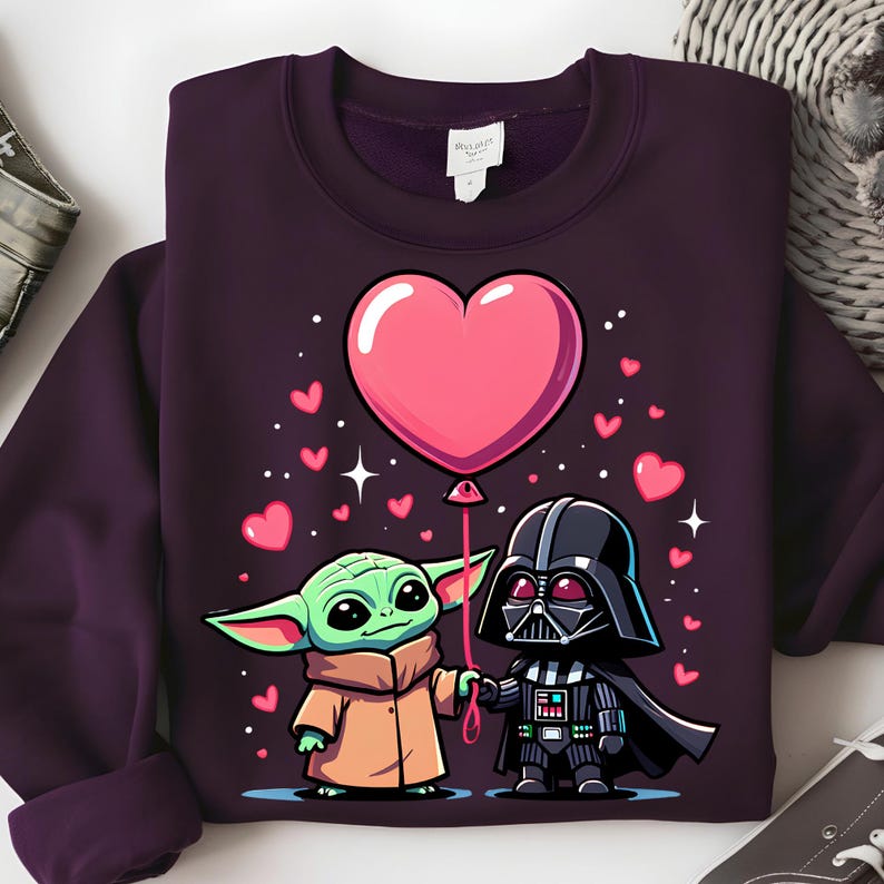 May include: A dark purple sweatshirt with a cartoon illustration of Baby Yoda and Darth Vader holding hands. A large pink heart balloon floats above them, surrounded by smaller pink hearts and stars. The sweatshirt is a solid colour.