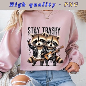 Puede incluir: Sudadera rosa con un gráfico de dos mapaches tocando rock and roll. Los mapaches llevan chaquetas de cuero y uno de ellos sostiene una guitarra. El texto "Stay Trashy" está impreso sobre los mapaches.