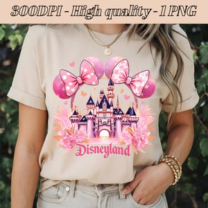 以下が含まれることがあります： ベージュのTシャツに、ピンクと白の城のグラフィックがプリントされています。城の下には「Disneyland」の文字が書かれています。グラフィックには、ピンクと白のミニーマウスの耳も含まれています。