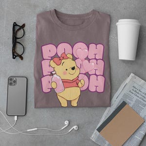May include: A brown t-shirt with a pink graphic of Winnie the Pooh holding a bottle and pink balloons. The text "POOH" is repeated in pink letters.